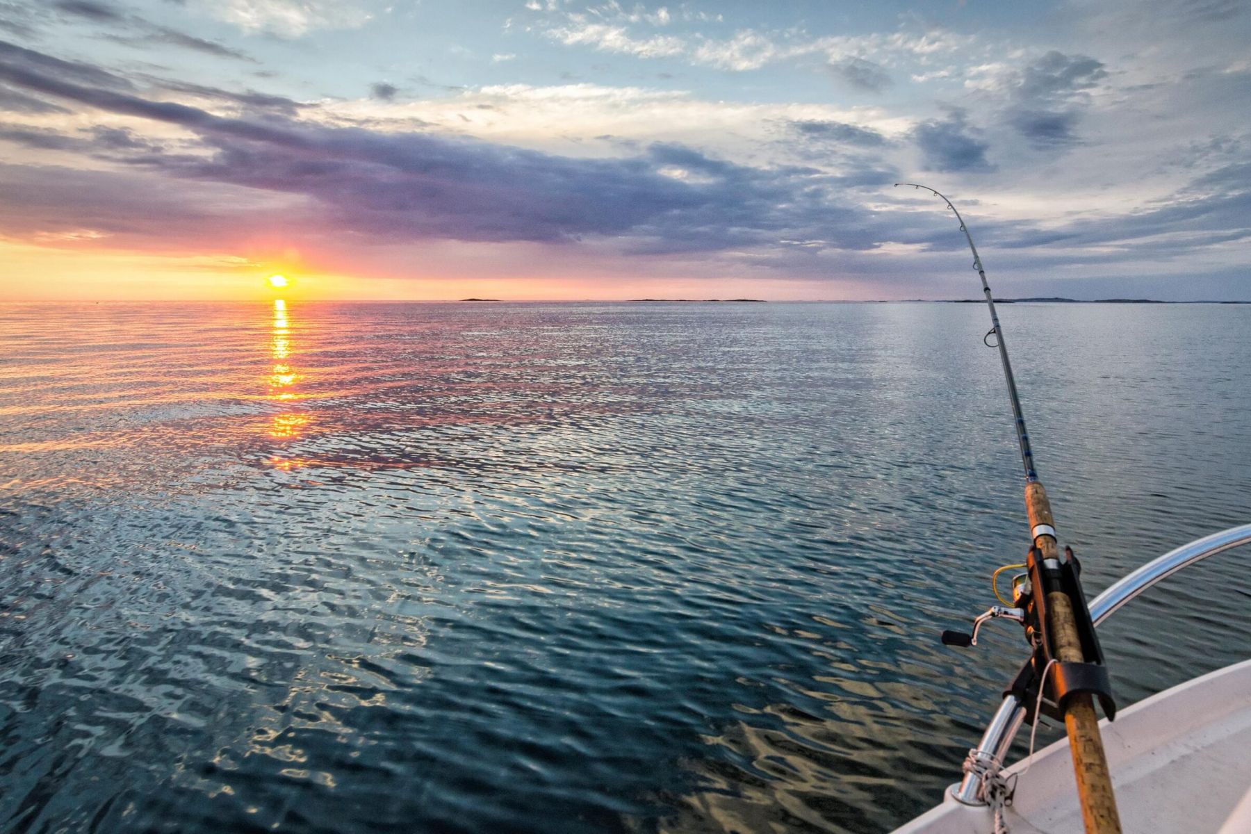 Person fishing from a boat on calm water at sunset, with colorful clouds and the sun near the horizon.