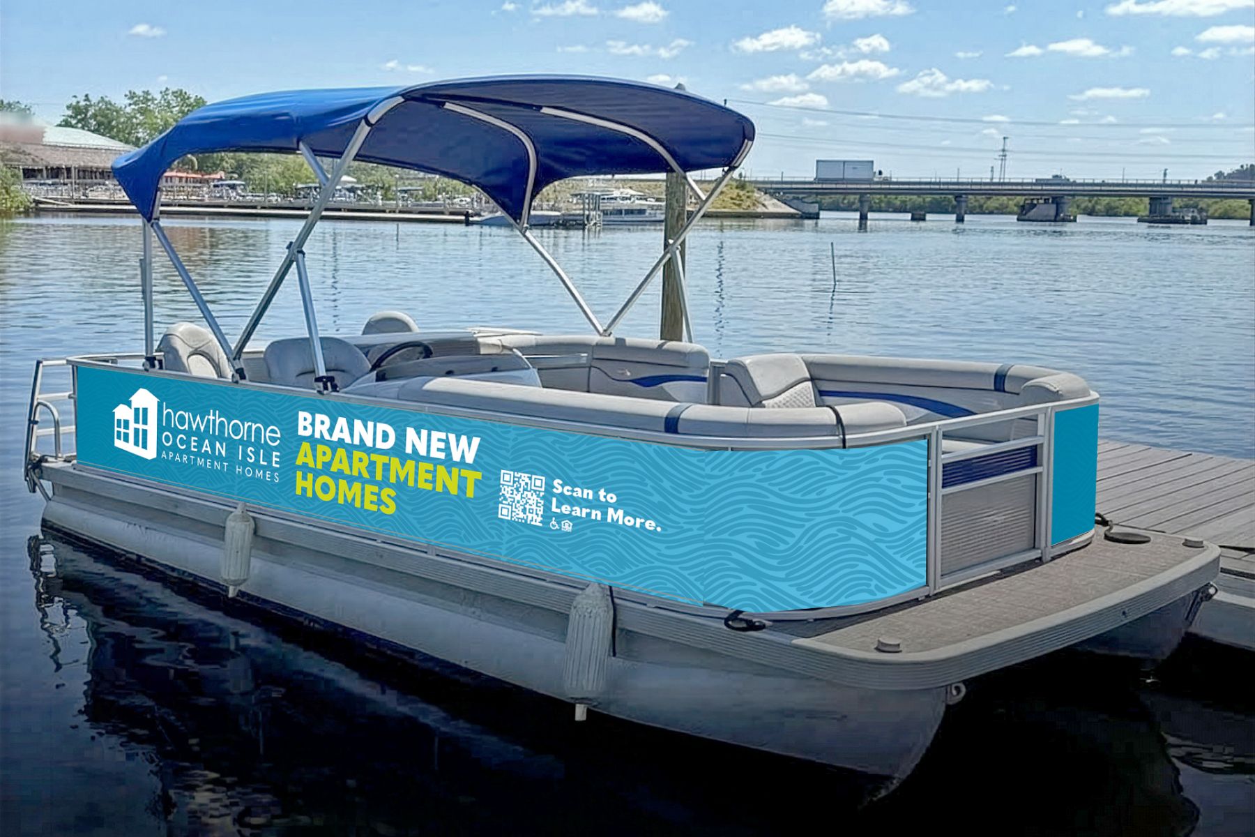 Blue and gray pontoon boat with a canopy, docked on a sunny day, displaying an ad for apartment homes.