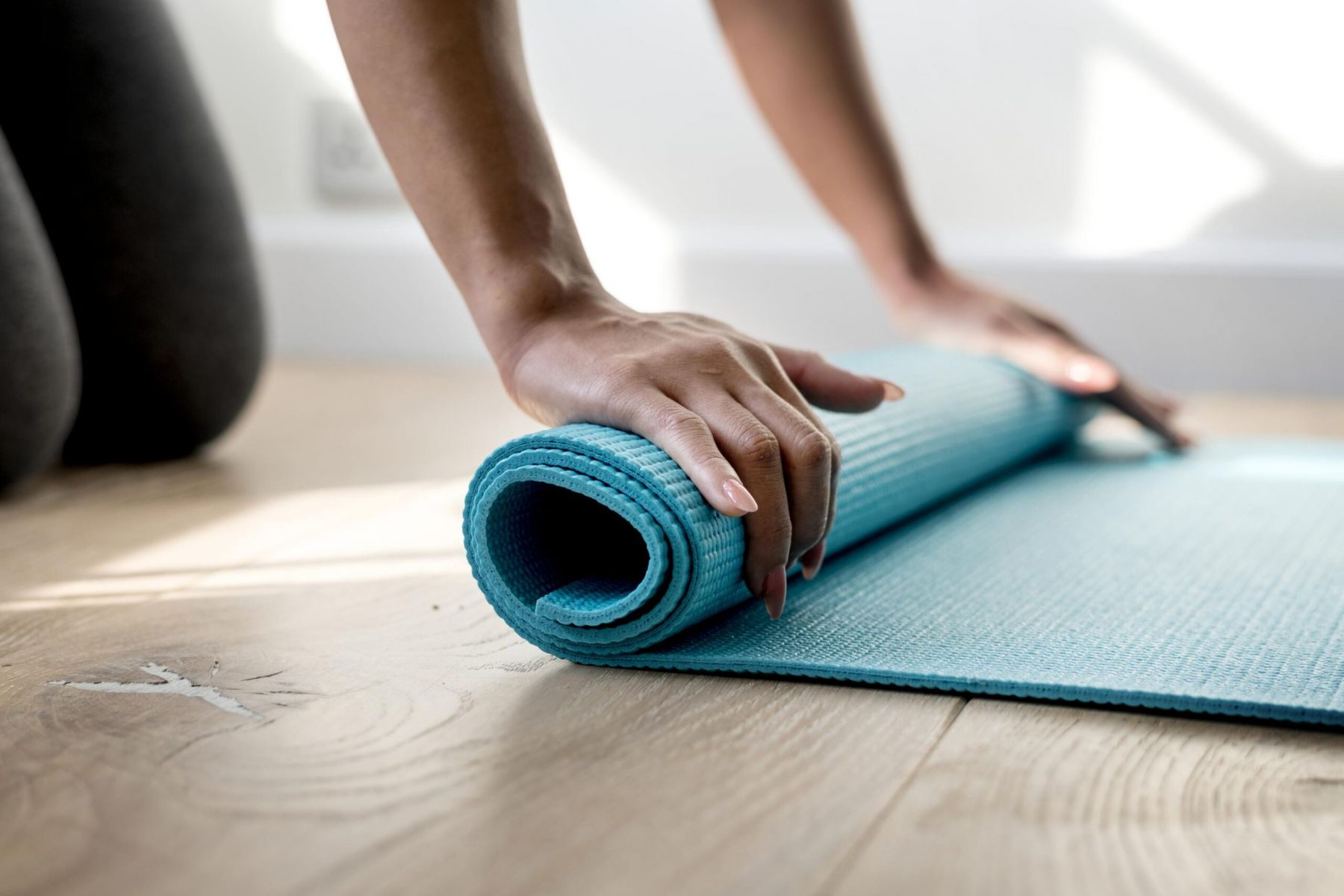 Close-up of hands rolling up a blue yoga mat on a wooden floor.