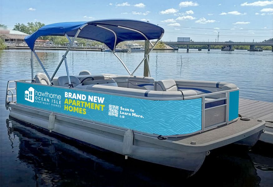 Blue and gray pontoon boat with a canopy, docked on a sunny day, displaying an ad for apartment homes.
